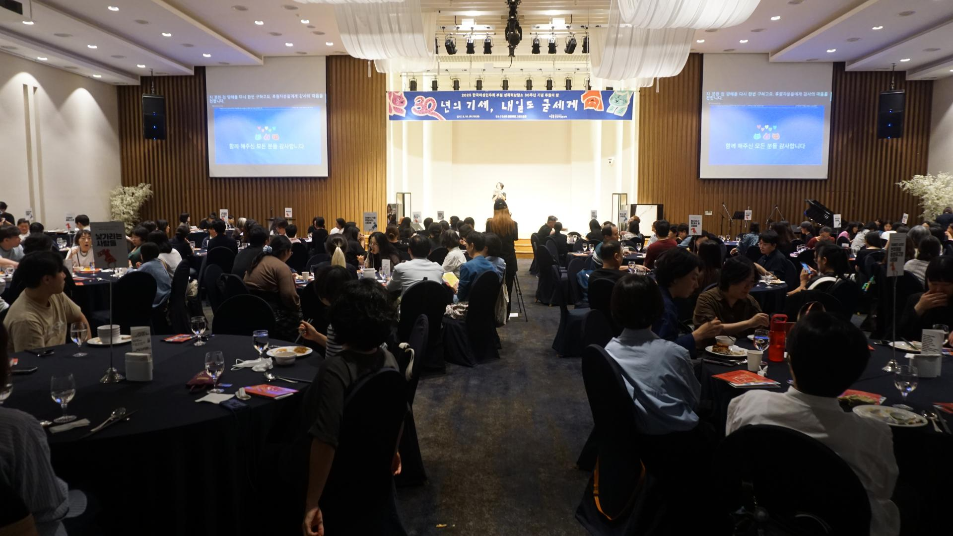 행사장의 모습. 원형 테이블에 앉은 사람들이 무대 앞쪽을 쳐다보고 있고, 무대에는 사회자가 나와 뭔가를 설명하고 있다. 양쪽 스크린에는 파란 화면에 '함께해주신 모든 분들 감사합니다'라고 쓰여 있다. 