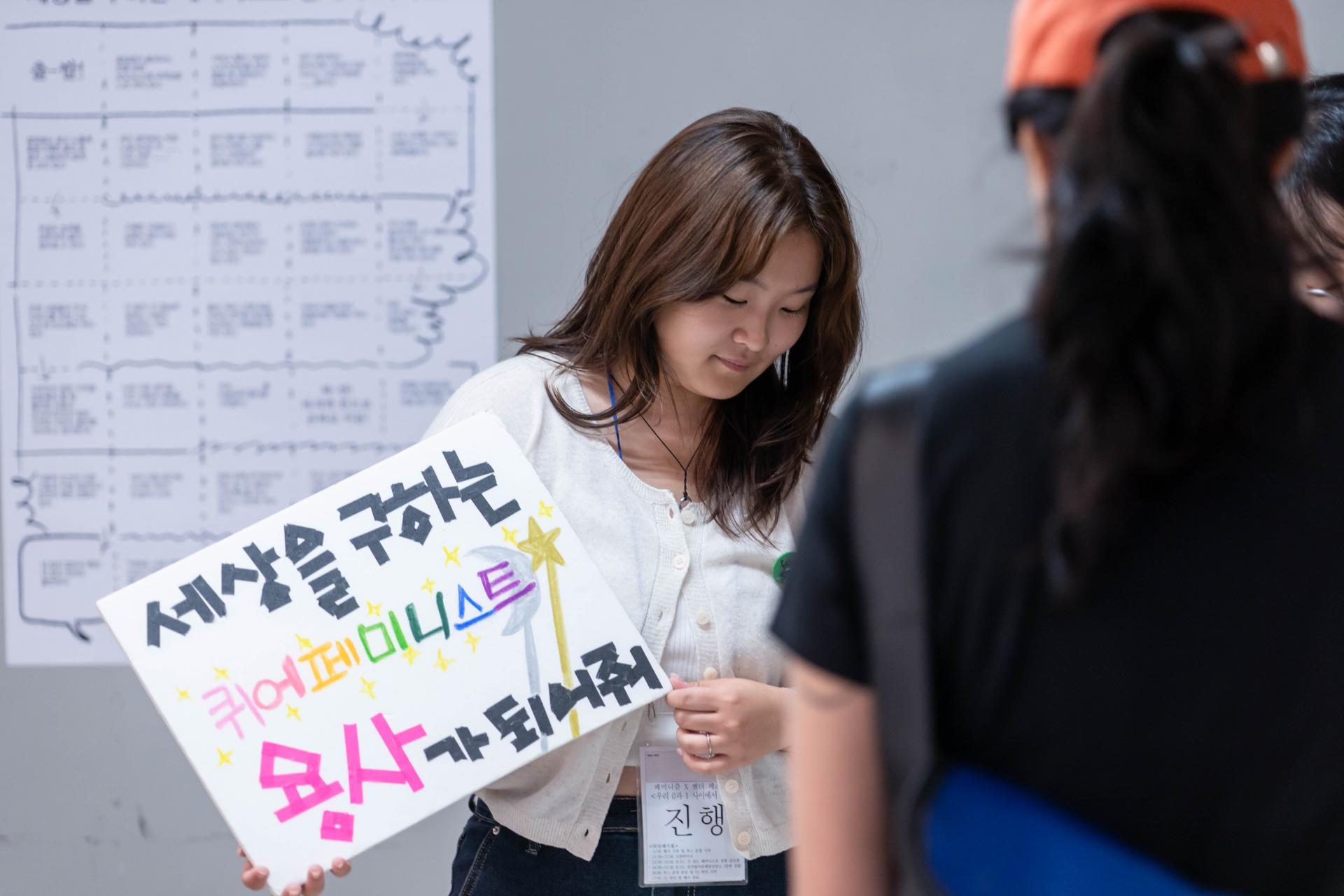 세상을 구하는 퀴어 페미니스트 용사가 되어줘가 적힌 판넬을 들고 있는 활동가의 모습과 프로그램에 참여하고 있는 참여자의 뒷모습