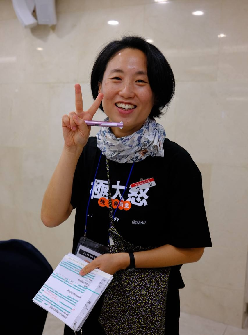 사회자가 무대가 아닌 일상적인 공간에서, 밑줄이 잔뜩 쳐진 대본노트와, 펜을 한 손에 들고, 펜을 든 손으로 브이를 그리며 카메라를 보고 웃고 있는 모습.