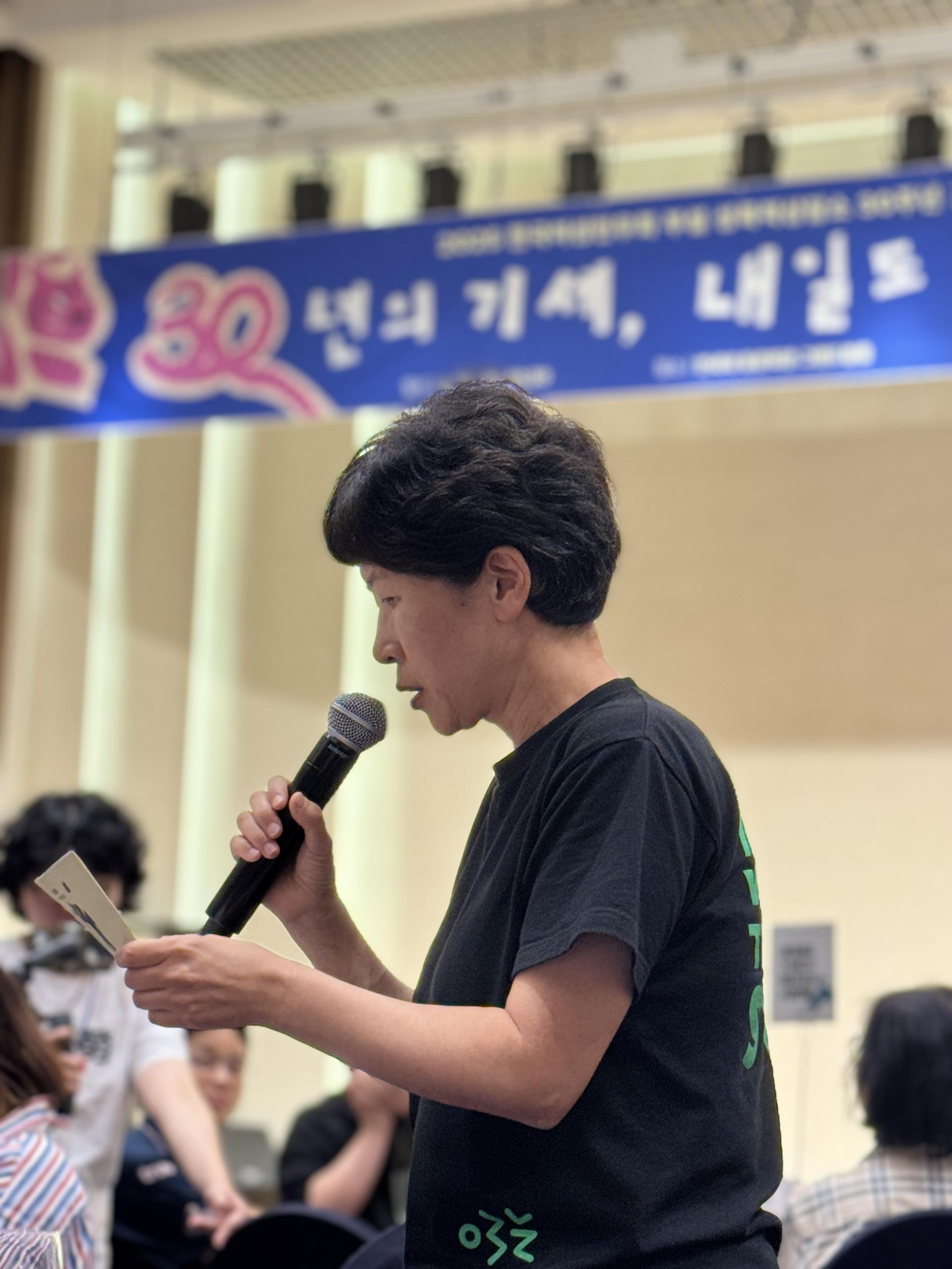 청중 사이 자리에서 일어나 발표하는 꼼꼼의 모습. 초록색 민우회 로고가 그려진 검은 티셔츠를 입고 있다. 마이크를 한 손에, 노트를 다른 한 손에 들고 발언하는 옆모습이다. 주변엔 앉아있는 다른청중들의 모습과 사진촬영중인 스탭의 모습이 흐릿하게 보인다.