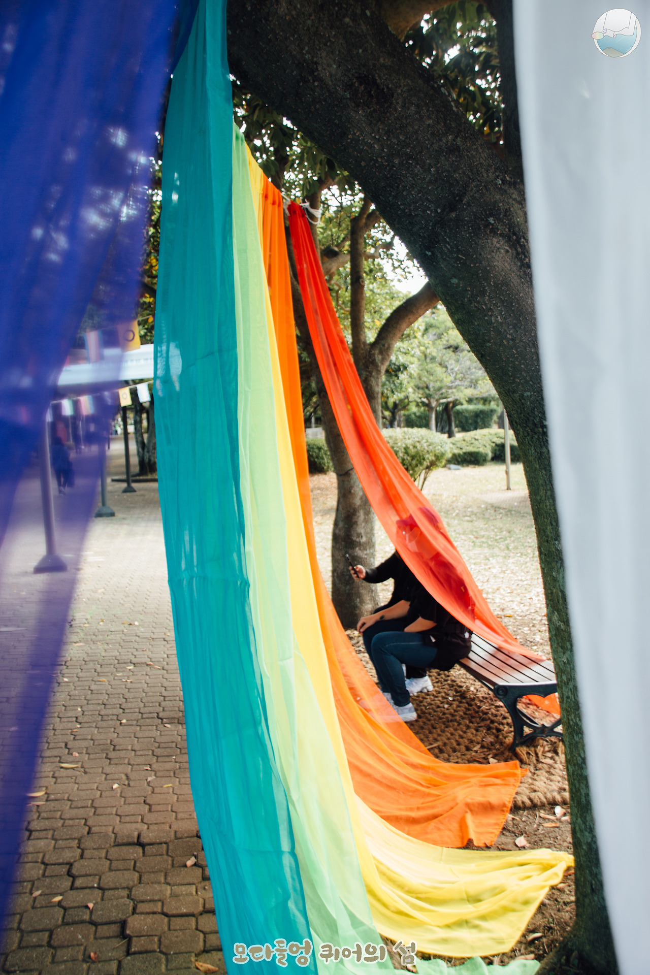 무지개 커튼을 걸치고 공원 벤치에 앉아 셀카를 촬영중인 자원활동가들의 모습