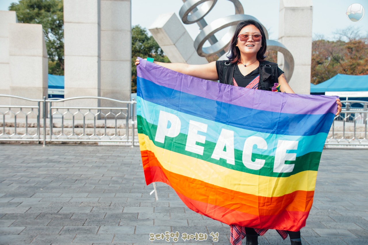 신산공원에서 하얀 글씨로 "PEACE"라고 적혀있는 무지개 깃발을 넓게 펼처들고 있는 혜영의 모습.