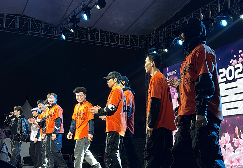 2025 동대문구 봄꽃축제 무대에서 갬블러크루(Gamblerz Crew) 댄서들이 오렌지색 유니폼을 입고 무대 위에 서 있는 모습. 조명이 반사된 무대 바닥과 벚꽃 이미지를 배경으로 한 LED 스크린이 봄 축제 분위기를 더한다.