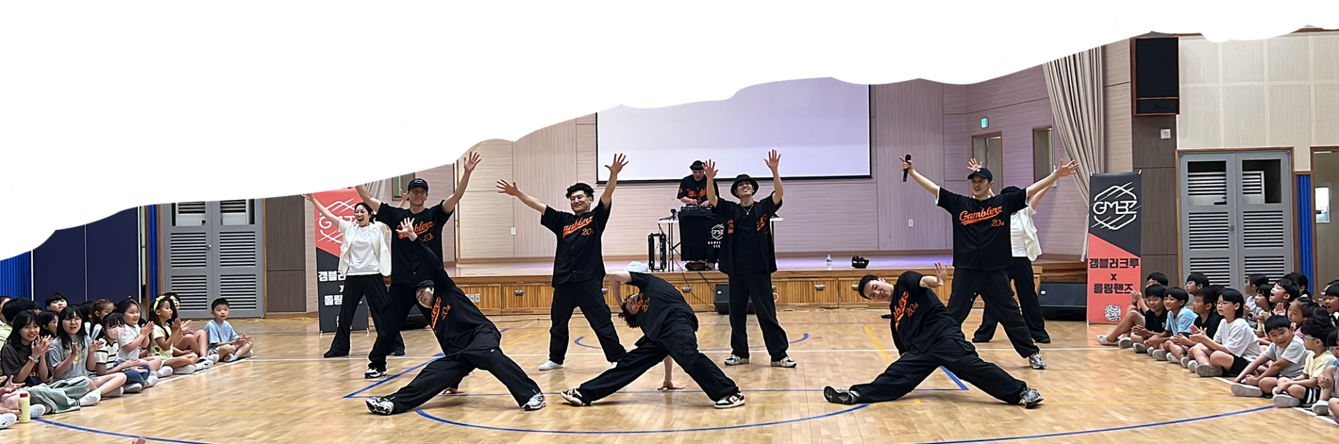 체육관 무대에서 갬블러크루가 초등학생 관객 앞에서 스트릿댄스를 선보이며 단체 포즈를 취하고 있는 모습