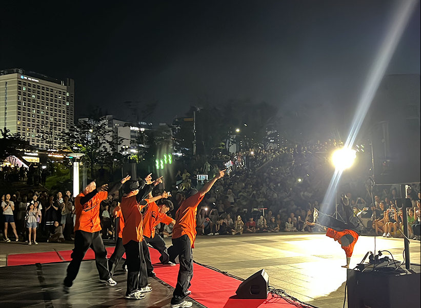 하남문화재단 스테이지 버스킹 무대에서 갬블러크루(Gamblerz Crew) 댄서들이 오렌지색 유니폼을 입고 야외 공연을 펼치는 장면. 밤하늘 아래 밝은 조명과 관객들의 환호 속에 스트릿댄스 퍼포먼스가 진행되고 있다.