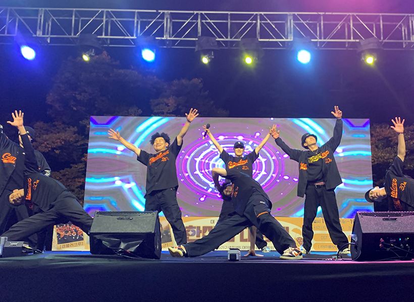 대전과학기술대학교 축제 무대에서 갬블러크루(Gamblerz Crew)가 검은색 의상에 오렌지 로고가 새겨진 유니폼을 입고 역동적인 스트릿댄스 퍼포먼스를 펼치는 장면. 무대 뒤 LED 스크린에는 화려한 그래픽이 연출되고, 조명 아래에서 댄서들의 에너지가 빛난다.