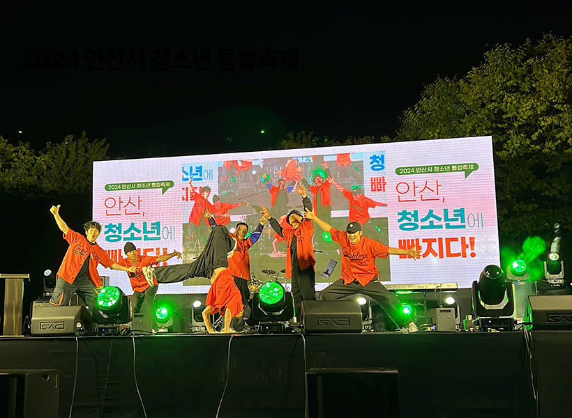 2024 안산시 청소년 통합축제 무대에서 갬블러크루(Gamblerz Crew)가 오렌지색 의상을 입고 브레이킹 퍼포먼스를 펼치는 장면. 무대 뒤 LED 스크린에는 ‘안산, 청소년에 빠지다!’라는 문구가 크게 비치고, 녹색 조명이 축제 분위기를 더한다.