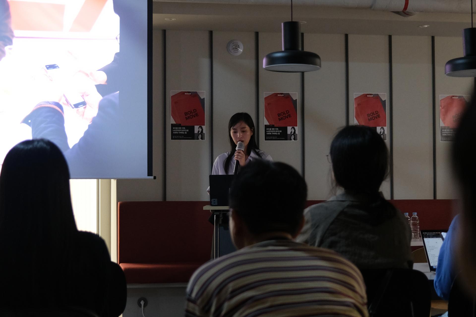 여성 발표자가 마이크를 들고 북토크에서 발표 중이고, 관객들이 경청하고 있다.