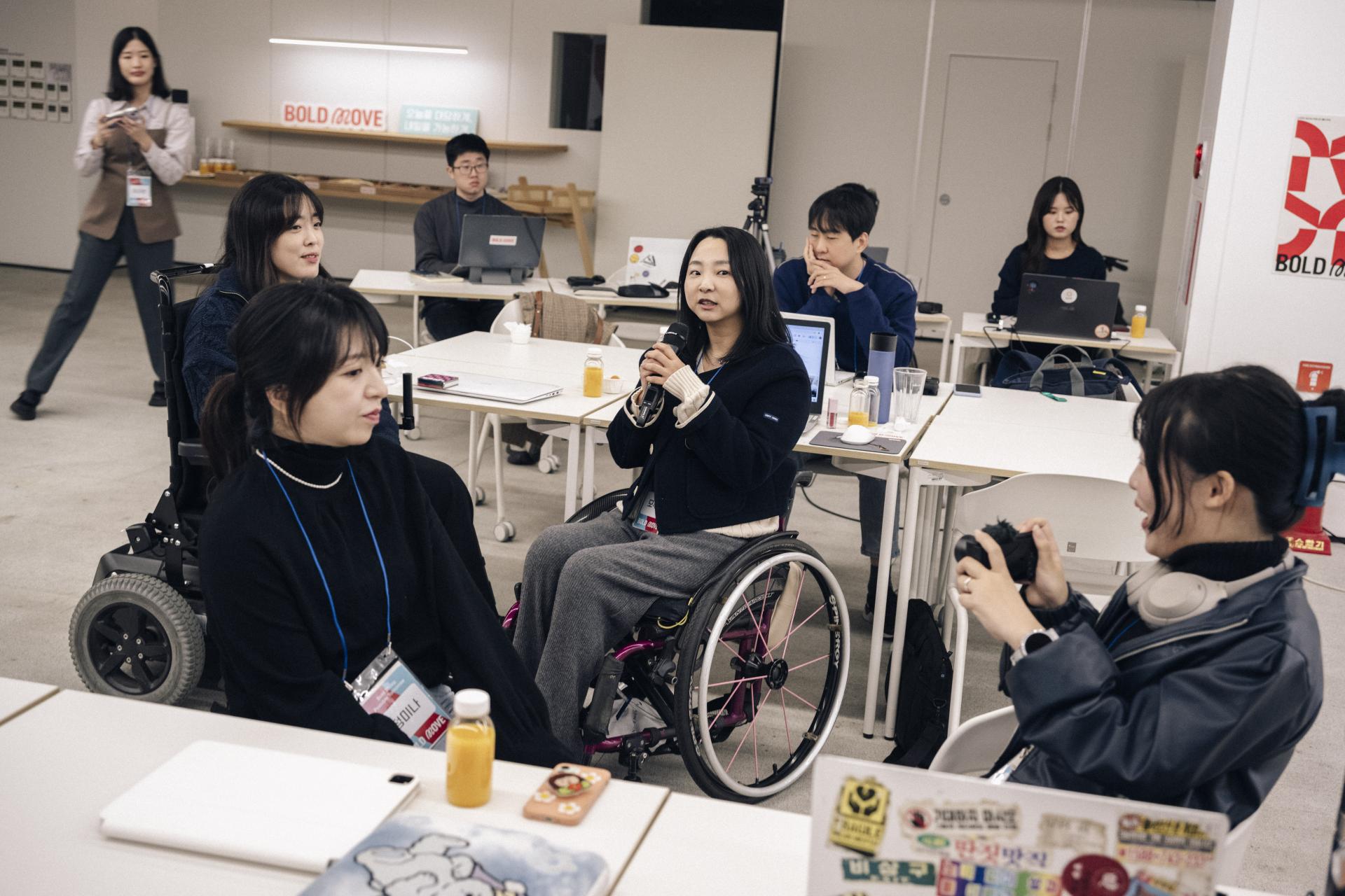 ‘BOLD MOVE’가 적힌 무대 앞에서 발표자가 마이크를 들고 이야기하고 있고, 멤버들이 소파에 앉아 발표를 듣고 있다.