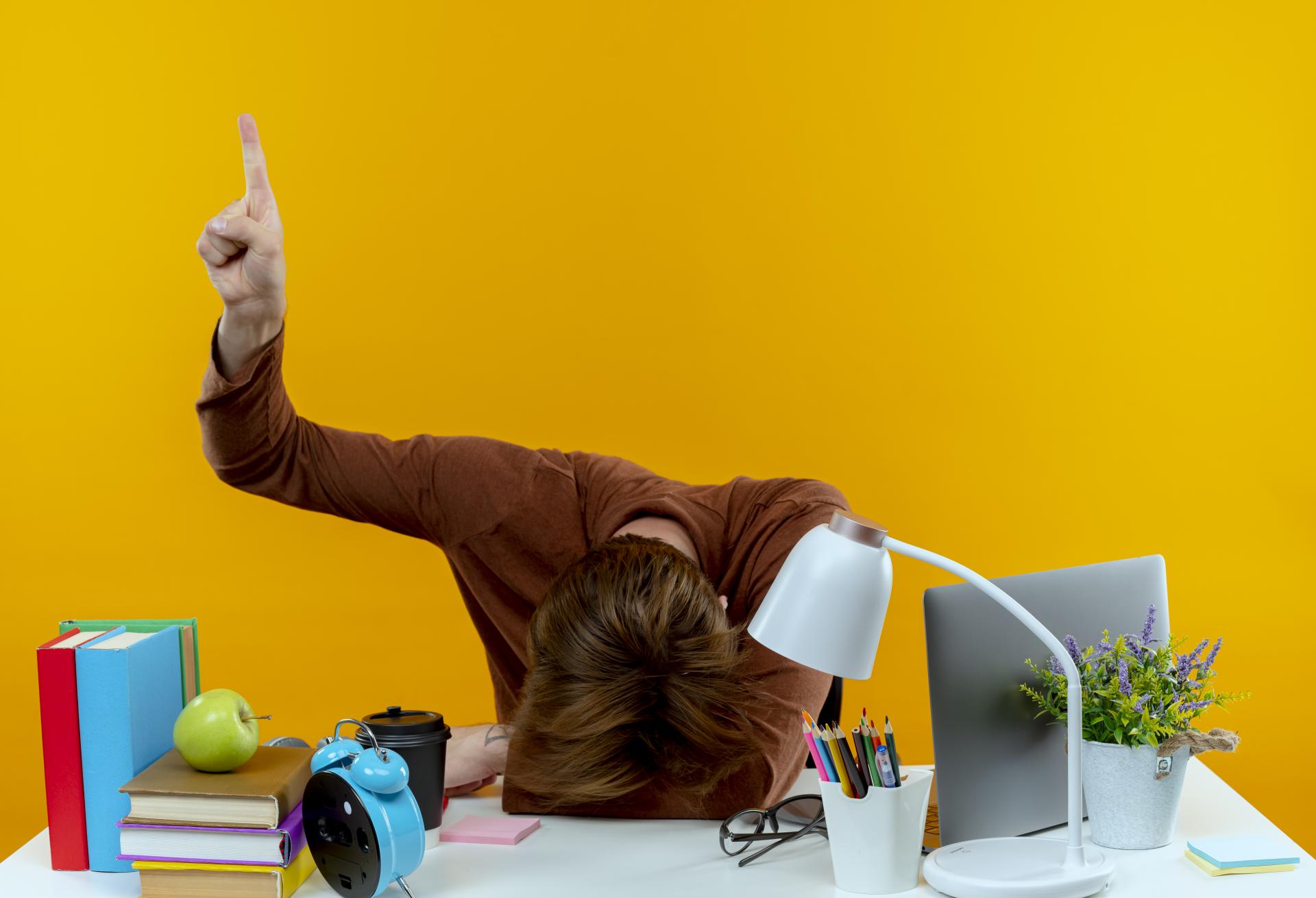 with-lowered-head-young-student-boy-sitting-desk-with-school-tools-points-up-isolated-yellow-wall-with-copy-space.jpg