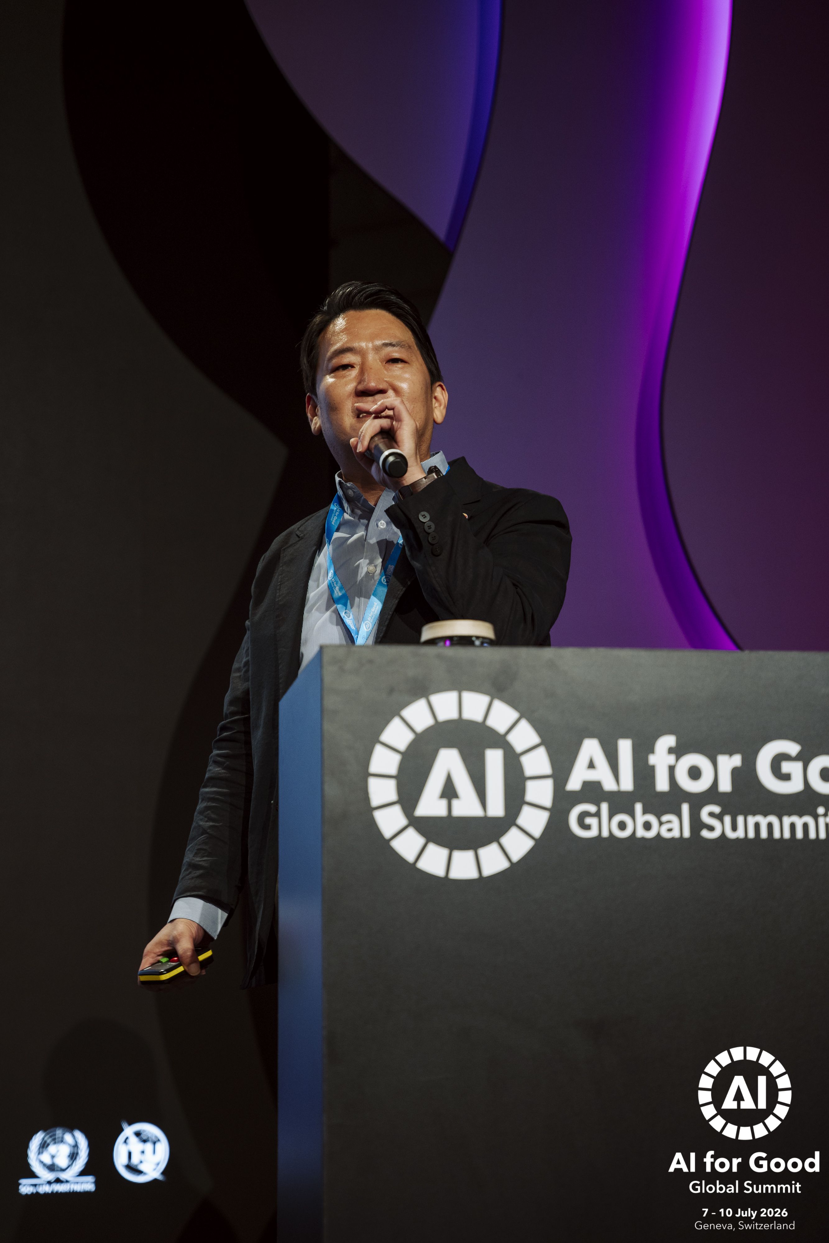 이큐포올 이인구 대표가 AI for Good Global Summit 2026 연단에서 마이크를 들고 발표하는 모습. 보라색 무대 조명 아래, 연단에 'AI for Good Global Summit' 로고가 보인다.
